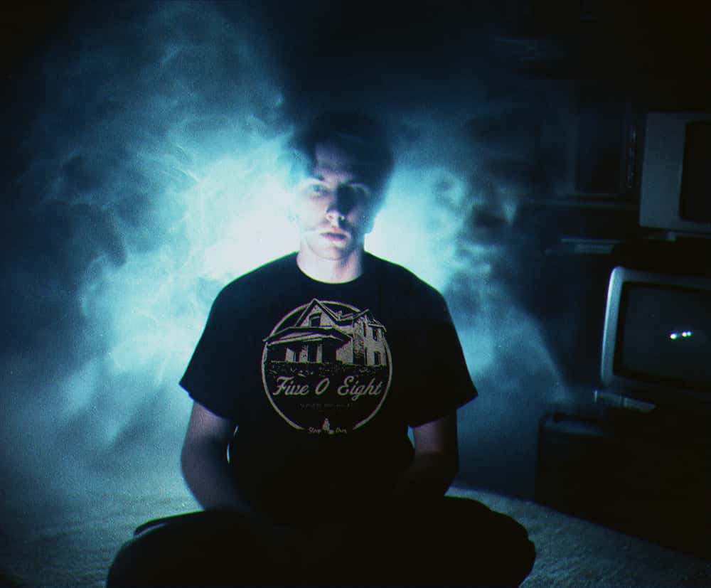 Model wearing SleepOver Clothing's Five O Eight haunted house t-shirt, sitting in a dark room with glowing ghostly mist behind them.