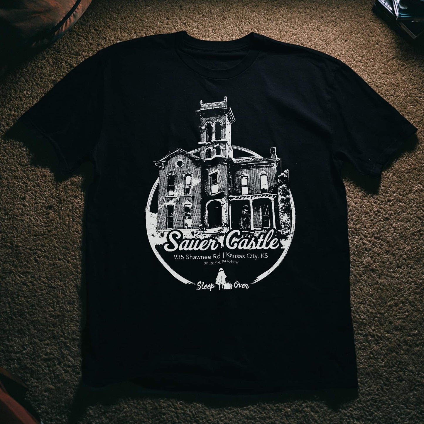 A black short-sleeve t-shirt laid flat on a textured brown carpet, featuring a large white graphic print of 'The Sauer Castle' in Kansas City, Kansas.