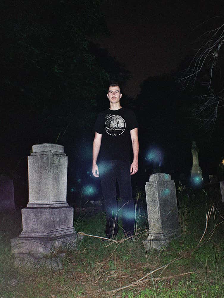 Model standing among old gravestones at night wearing SleepOver Clothing’s Stull Cemetery haunted location t-shirt, with eerie blue orbs glowing around them.