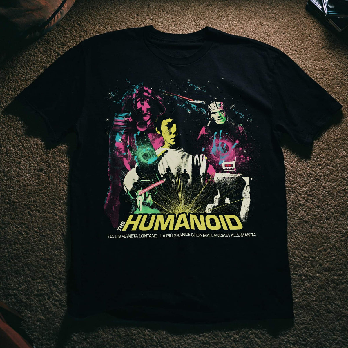 A black graphic t-shirt featuring a colorful sci-fi design of the character 'The Humanoid' with neon green, pink, and yellow details, laid flat on a brown carpet.