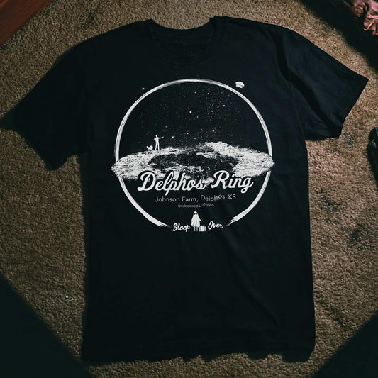 Black T-shirt featuring a white illustration of the Delphos Ring—an alleged UFO landing site on Johnson Farm in Delphos, Kansas—set beneath a starlit sky with figure and dog silhouettes. Text includes “Undisclosed Location” and “Sleep Over” branding.