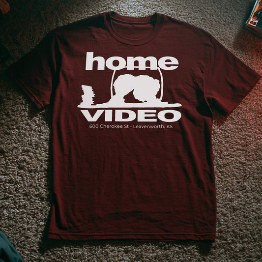 Maroon T-shirt with a retro-style white logo reading “Home Video” and an outline of a person crouched near a TV screen, with text “600 Cherokee St • Leavenworth, KS.”
