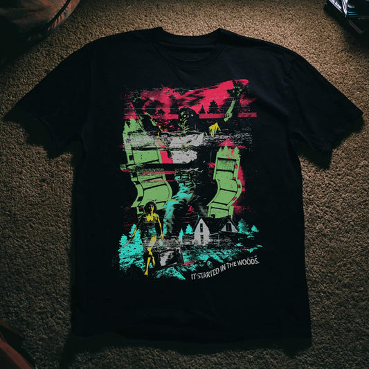 Black t-shirt with a glitchy, VHS-style multicolor collage design in pink, green, and teal, featuring a masked figure, a woman with a knife, a wooded cabin, and a television, with the tagline 'It Started in the Woods' below.