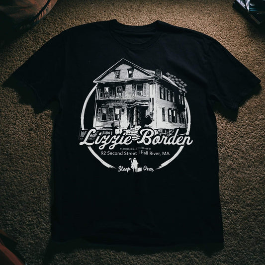 A black short-sleeve graphic t-shirt laid flat on a textured brown carpet. The shirt features a white circular graphic of a two-story house with the text 'Lizzie Borden' and 92 Second Street | Fall River, MA' printed on the front.