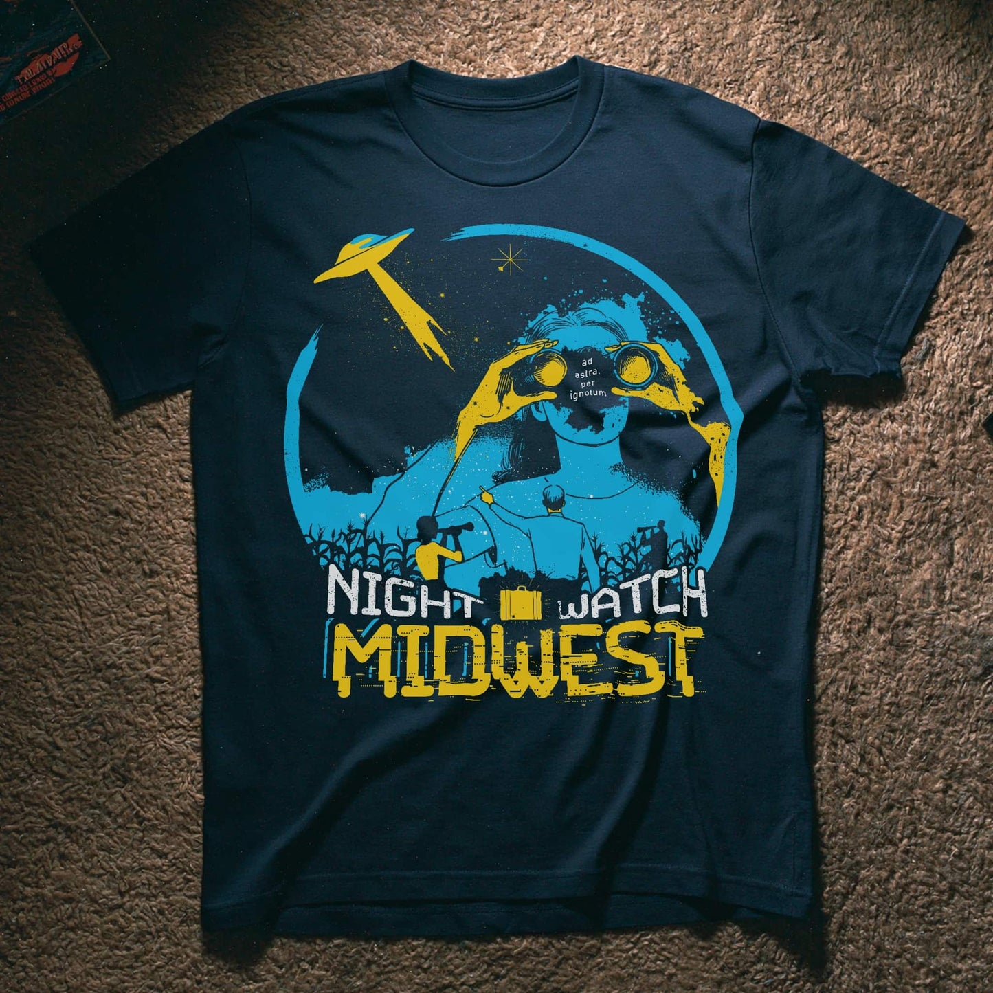 A dark navy blue graphic t-shirt laid flat on a brown carpet. The shirt features a distressed graphic of a figure with binoculars under a crescent moon and stars, with a yellow UFO, and the text 'NIGHT WATCH MIDWEST' in yellow and white.