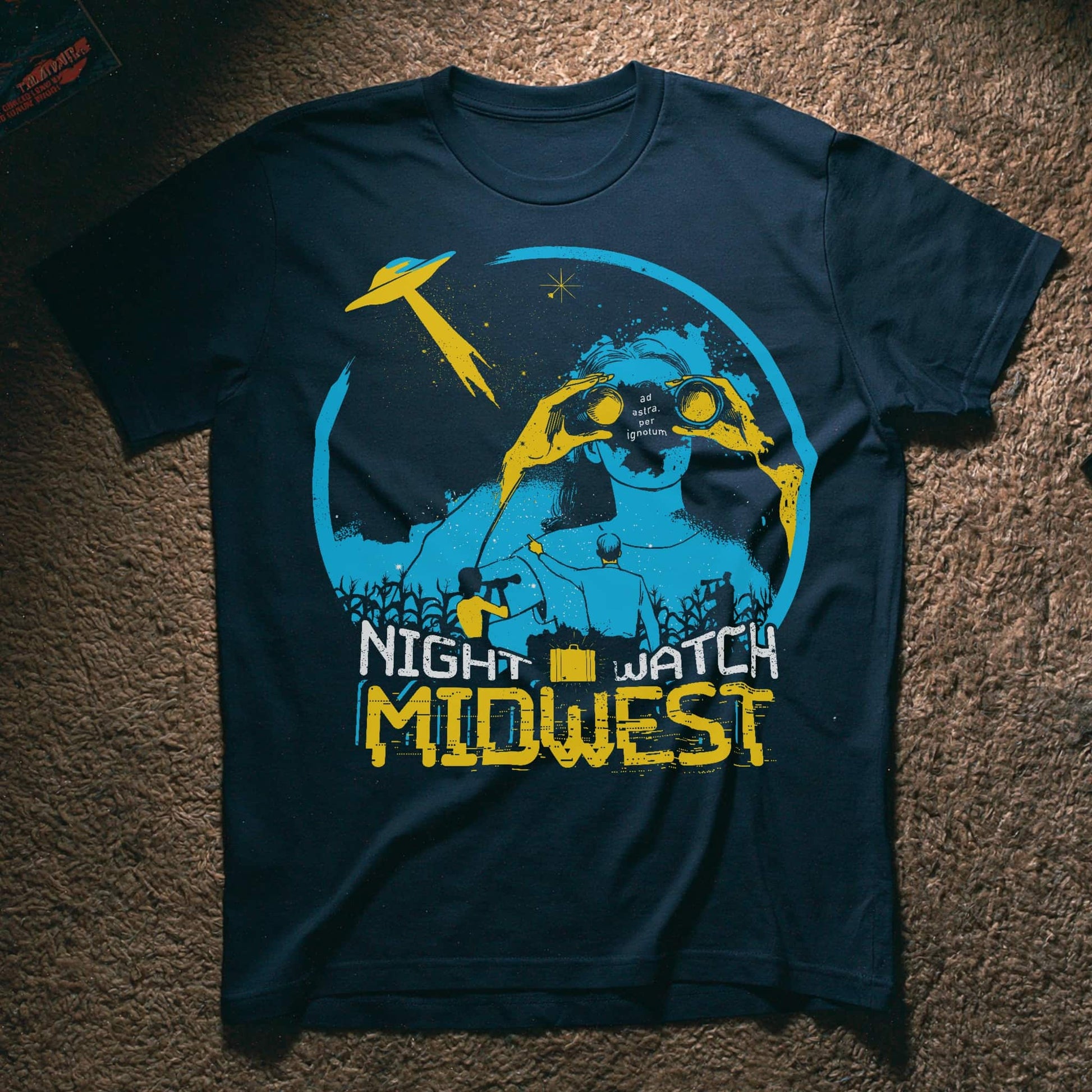A dark navy blue graphic t-shirt laid flat on a brown carpet. The shirt features a distressed graphic of a figure with binoculars under a crescent moon and stars, with a yellow UFO, and the text 'NIGHT WATCH MIDWEST' in yellow and white.