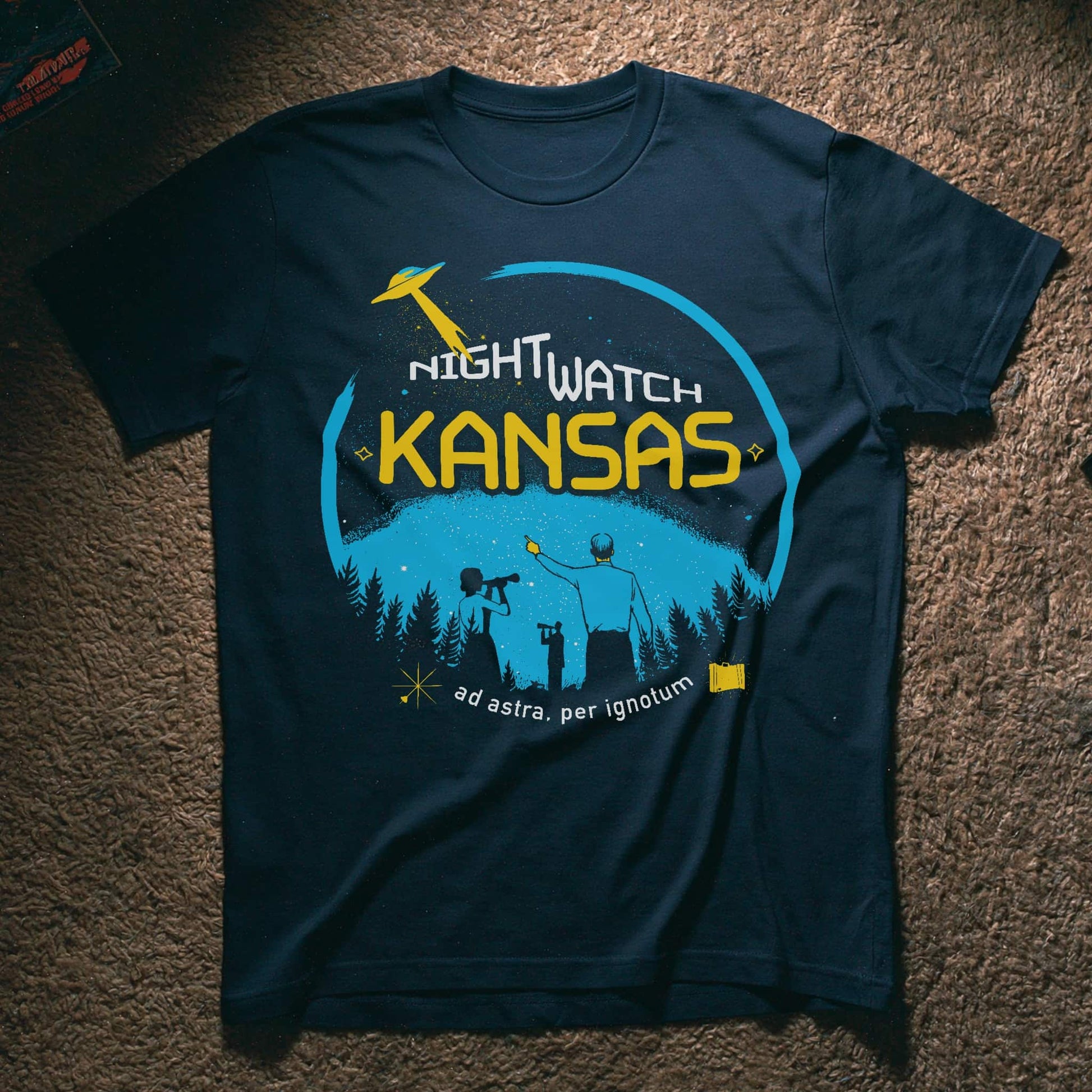 A navy blue graphic t-shirt laid flat on a brown carpet. The shirt features a circular graphic with a yellow UFO, the words 'NIGHTWATCH KANSAS' in white and yellow, and silhouettes of people looking up at a starry night sky above a pine tree line.