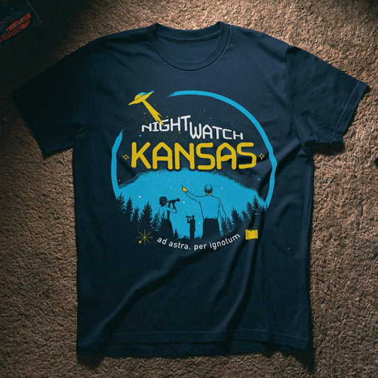 A navy blue graphic t-shirt laid flat on a brown carpet. The shirt features a circular graphic with a yellow UFO, the words 'NIGHTWATCH KANSAS' in white and yellow, and silhouettes of people looking up at a starry night sky above a pine tree line.