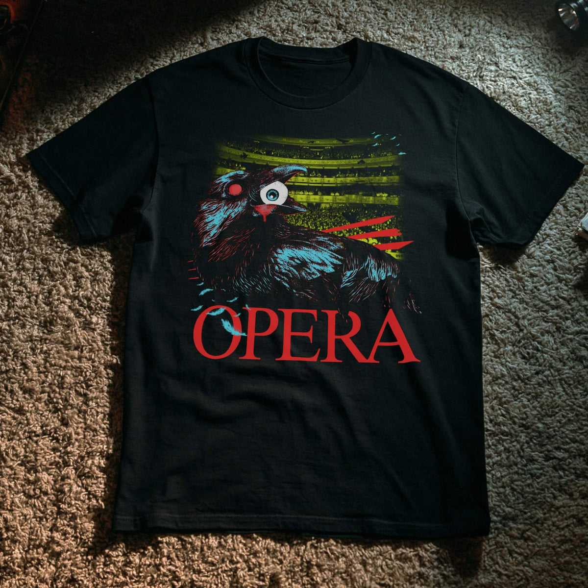 A black short-sleeve graphic t-shirt laid flat on a brown carpet. The shirt features a stylized raven with a human eye, blue and green feather details, red scratch-like accents, and the word 'OPERA' printed in red below.