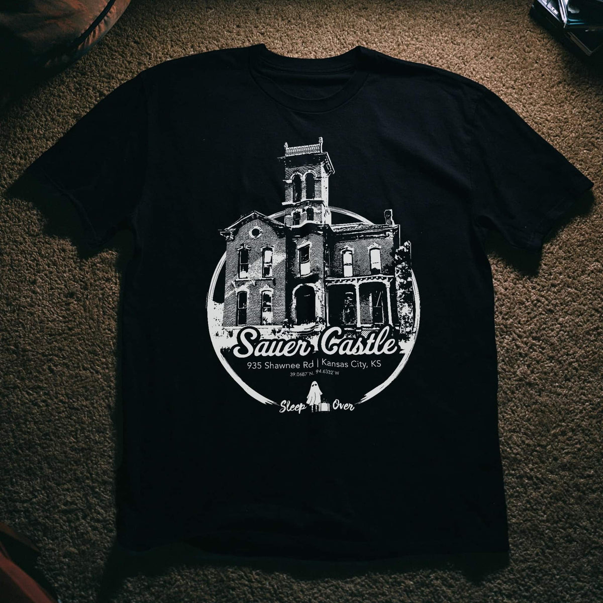 A black short-sleeve t-shirt laid flat on a textured brown carpet, featuring a large white graphic print of 'The Sauer Castle' in Kansas City, Kansas.