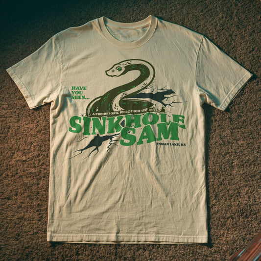 Cream-colored T-shirt with a green snake-like cryptid emerging from cracked earth, with the text ‘Sinkhole Sam – A Prehistoric Relic from the Deep, Inman Lake, KS.’