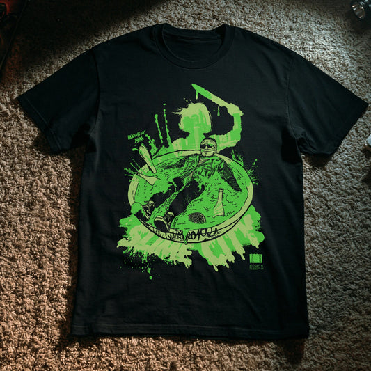 A black short-sleeve graphic t-shirt laid flat on a brown carpet. The shirt features a large neon green and white graphic print on the front depicting a character in a bowl with the word 'Slime City' and a small green Suitcase logo in the bottom right corner.