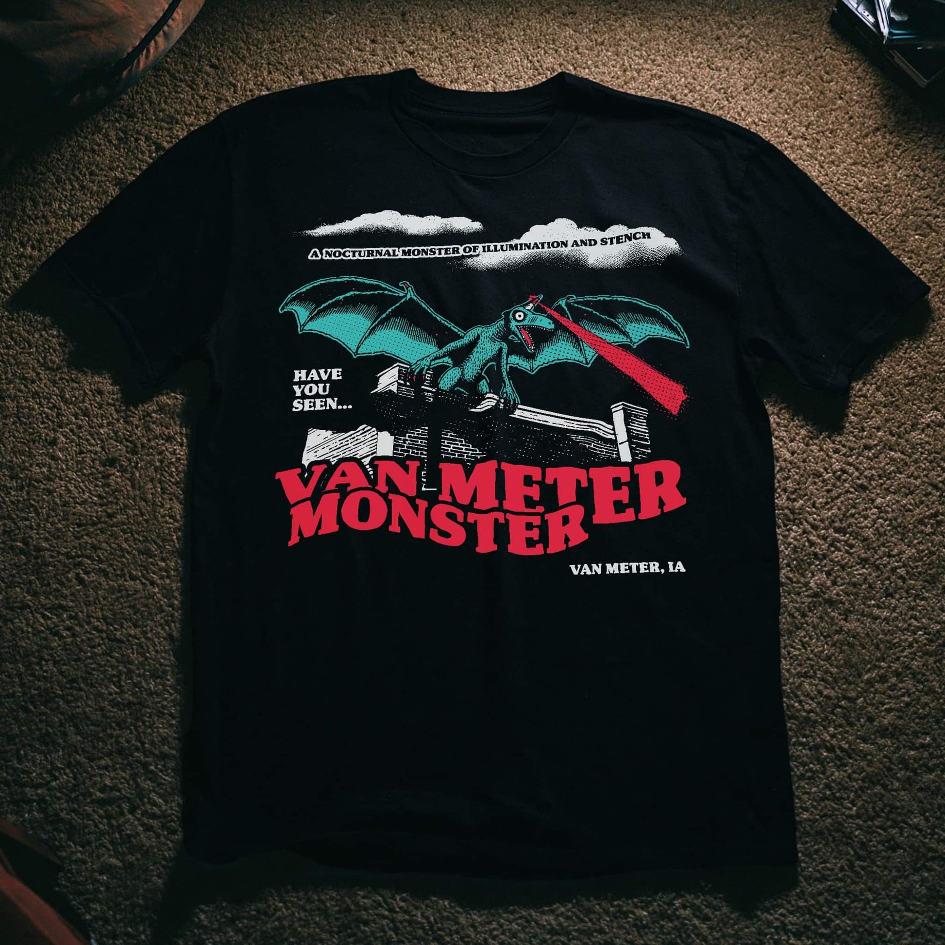 Black t-shirt with an illustration of a winged, dragon-like creature shooting a red light beam from its forehead, perched on a brick wall, with text reading 'Have You Seen... Van Meter Monster, Van Meter, IA' and 'A Nocturnal Monster of Illumination and Stench.