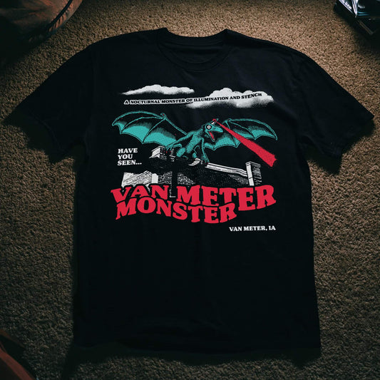 Black t-shirt with an illustration of a winged, dragon-like creature shooting a red light beam from its forehead, perched on a brick wall, with text reading 'Have You Seen... Van Meter Monster, Van Meter, IA' and 'A Nocturnal Monster of Illumination and Stench.