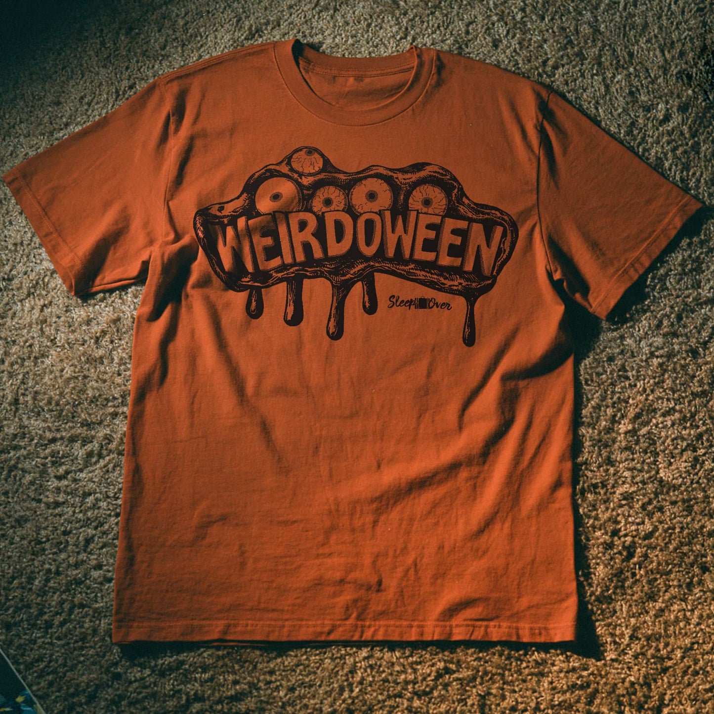 An orange crewneck t-shirt laid flat on a textured carpet background, featuring a dark brown graphic of a dripping brain with eyes and the word 'WEIRDOWEEN' across the chest.