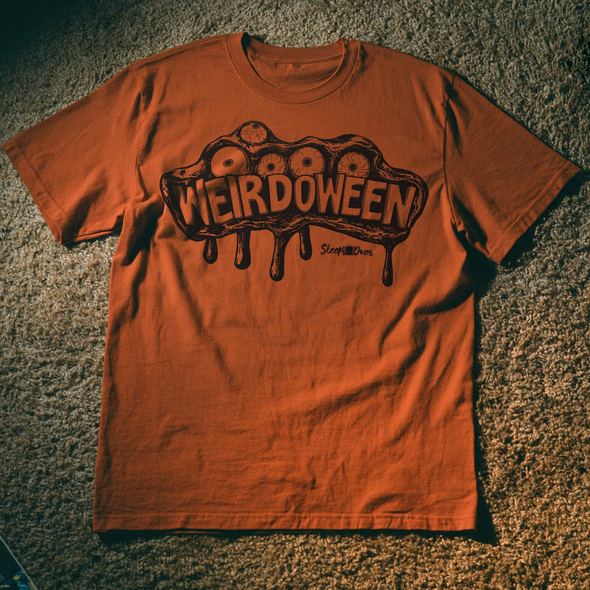 An orange crewneck t-shirt laid flat on a textured carpet background, featuring a dark brown graphic of a dripping brain with eyes and the word 'WEIRDOWEEN' across the chest.