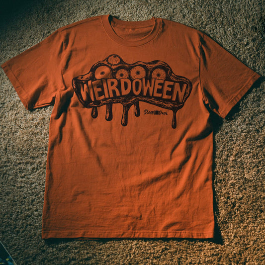 An orange crewneck t-shirt laid flat on a textured carpet background, featuring a dark brown graphic of a dripping brain with eyes and the word 'WEIRDOWEEN' across the chest.