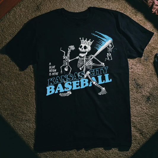 Black T-shirt featuring skeletons playing baseball, one with a crown and bat trailing a streak of blue, with the text ‘Kansas City Baseball’ and ‘A Blue Reign is Here.’