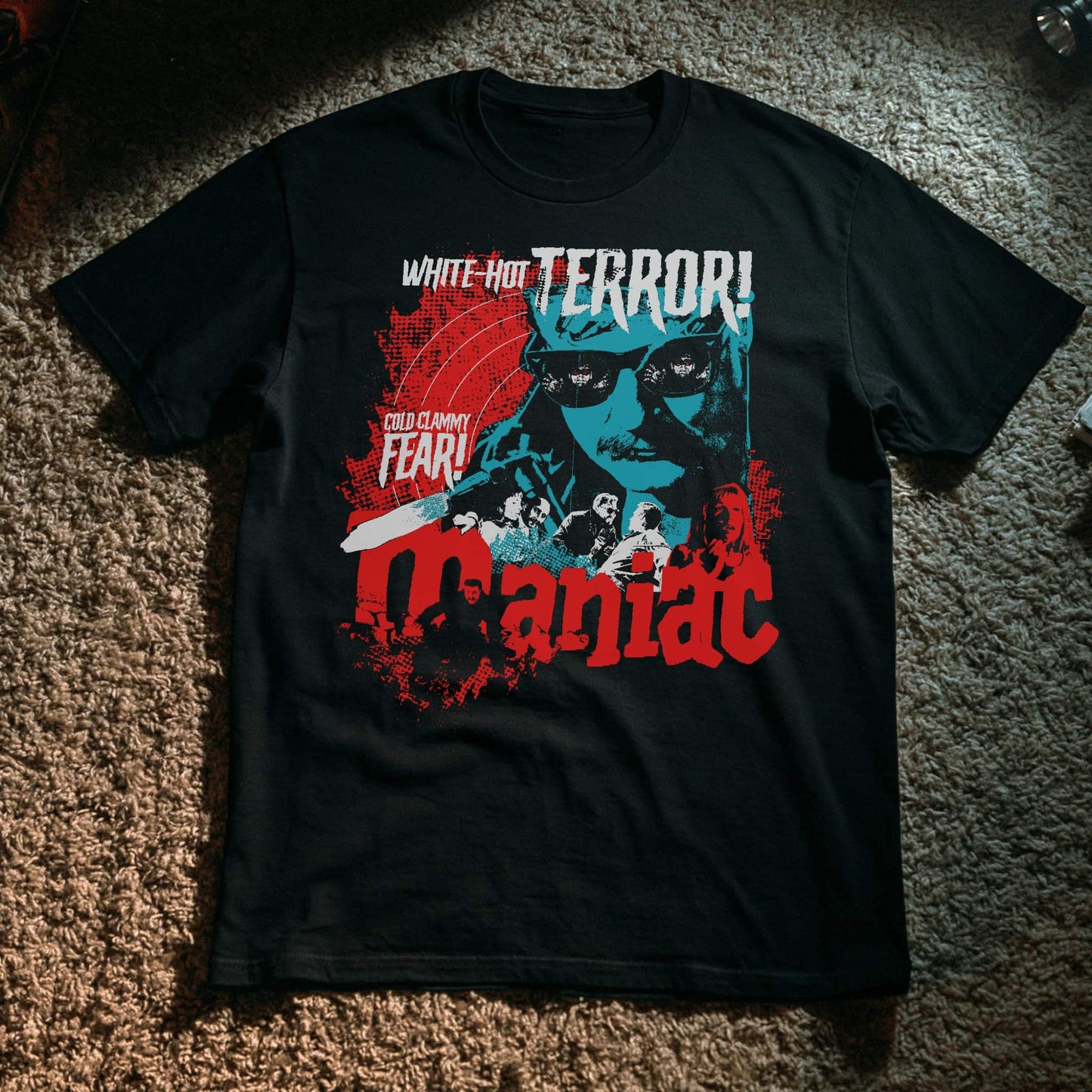 Black t-shirt with a vintage horror movie poster design in red, teal, and white for 'Maniac,' featuring a menacing man in sunglasses, a chainsaw, and a group of victims, with tagline text reading 'White-Hot Terror!' and 'Cold Clammy Fear!